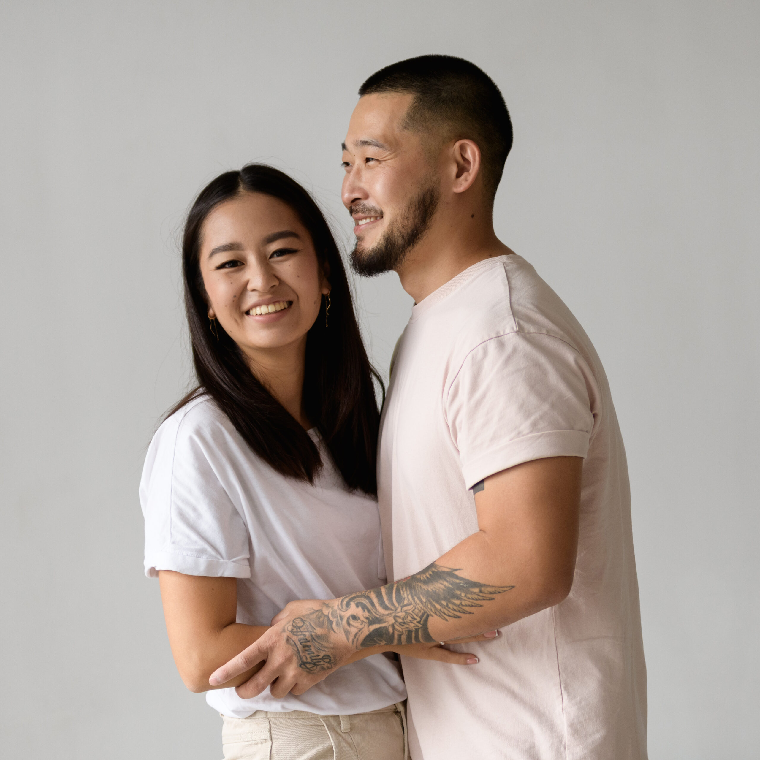Smiling asian woman hugging tattooed husband isolated on grey background