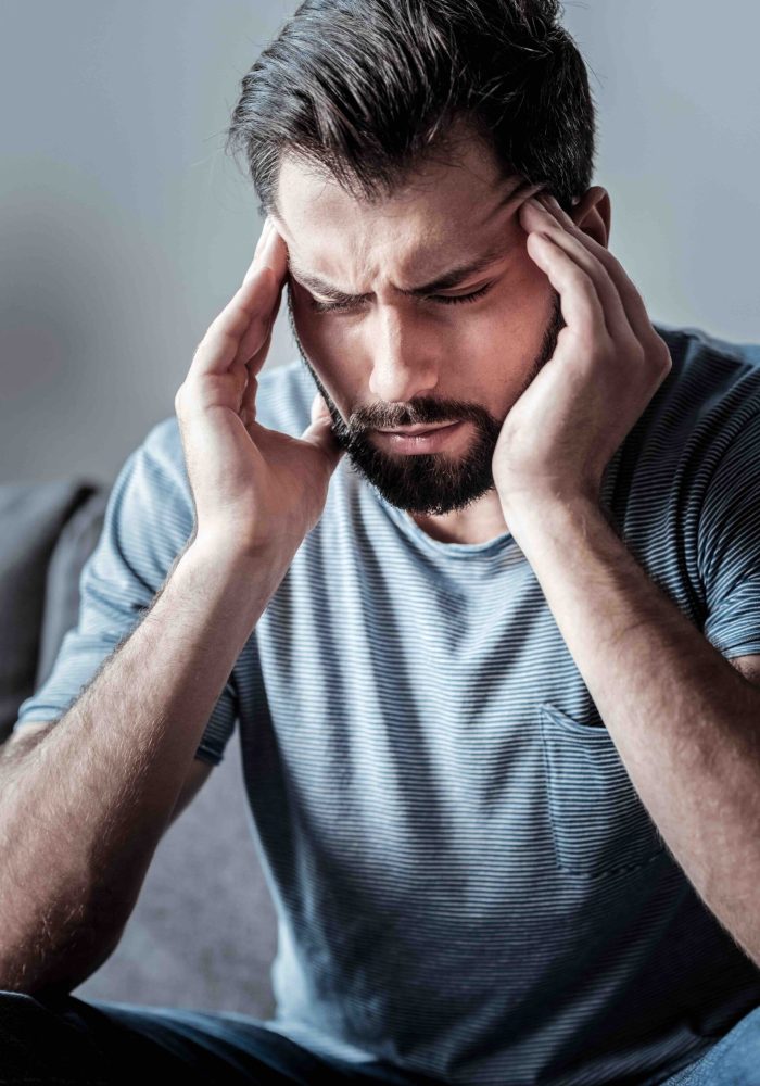 Strong headache. Sad nice young man sitting on the sofa and holding his temples while feeling headache