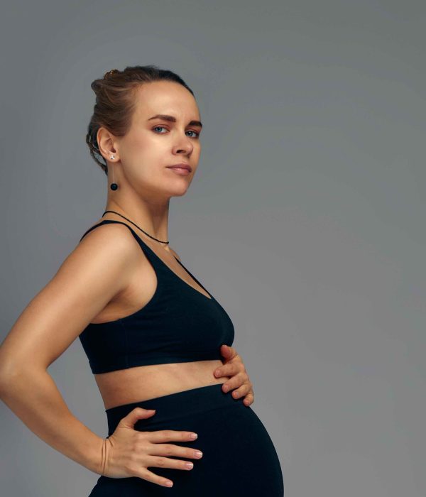 Cute pregnant belly isolated on gray. Side view of young pregnant woman embracing her abdomen with hands. Big belly on the third trimester of pregnancy close-up. Concept of pregnant life