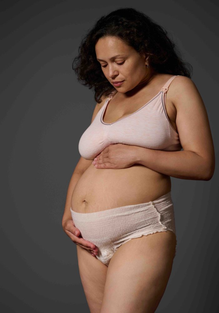 Studio portrait of new mom, a multi ethnic middle aged woman, touching her belly, isolated on gray background, dressed in lingerie and showing her body with stretch marks and postpartum flaws