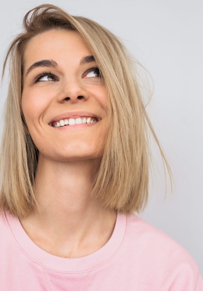 Closeup portrait of cheerful young blonde Caucasian female looking up with dreamy face wearing pink casual clothes, smiling and looking happy. Positive woman with healthy teeth smile with copy space.